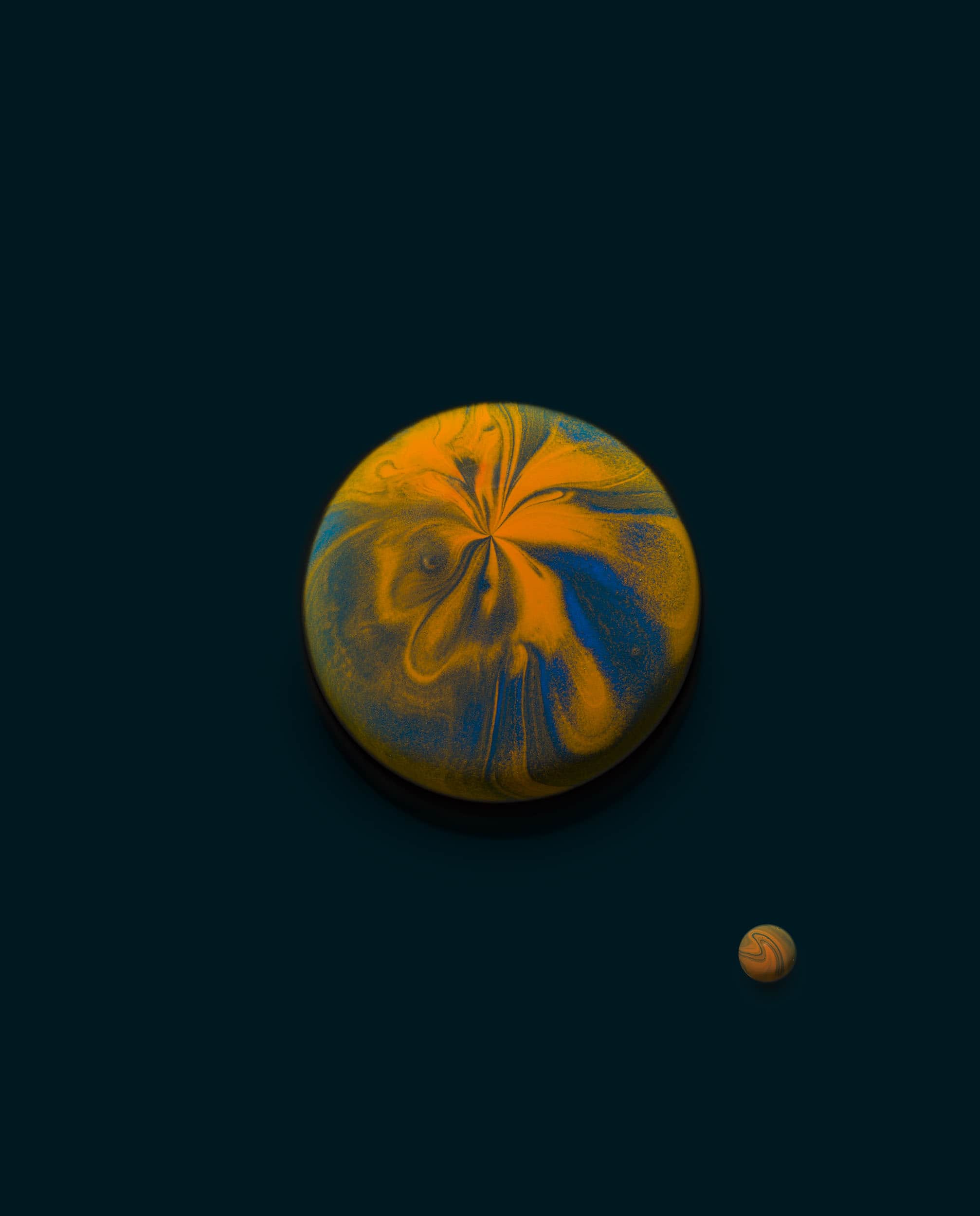 A mixture of orange and blue paint creating a marbled effect is shaped into a large spherical bubble, with a smaller one to accompany, on a black background. This looks like a planet with a moon orbiting it.