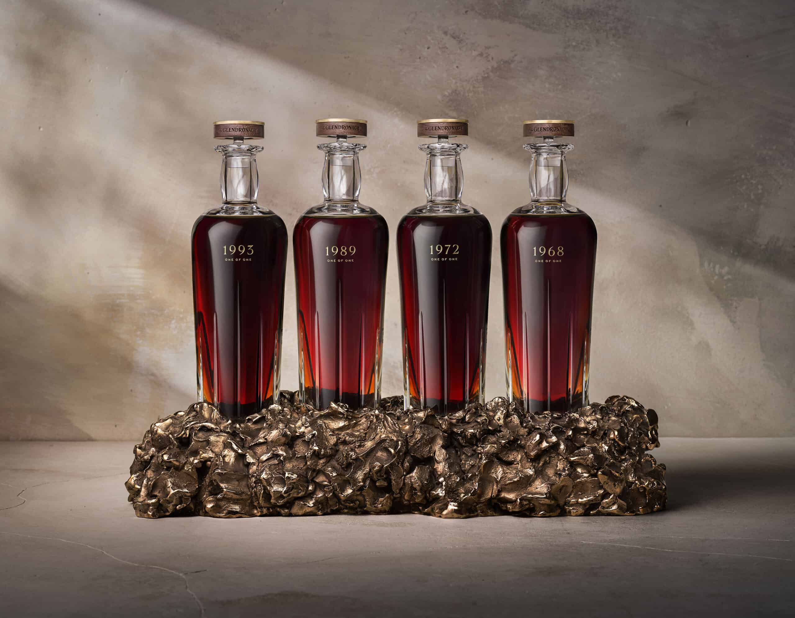 Four bottles of whisky sitting in a bronze sculpture on a neutral background. A single shaft of light highlights the bottles.