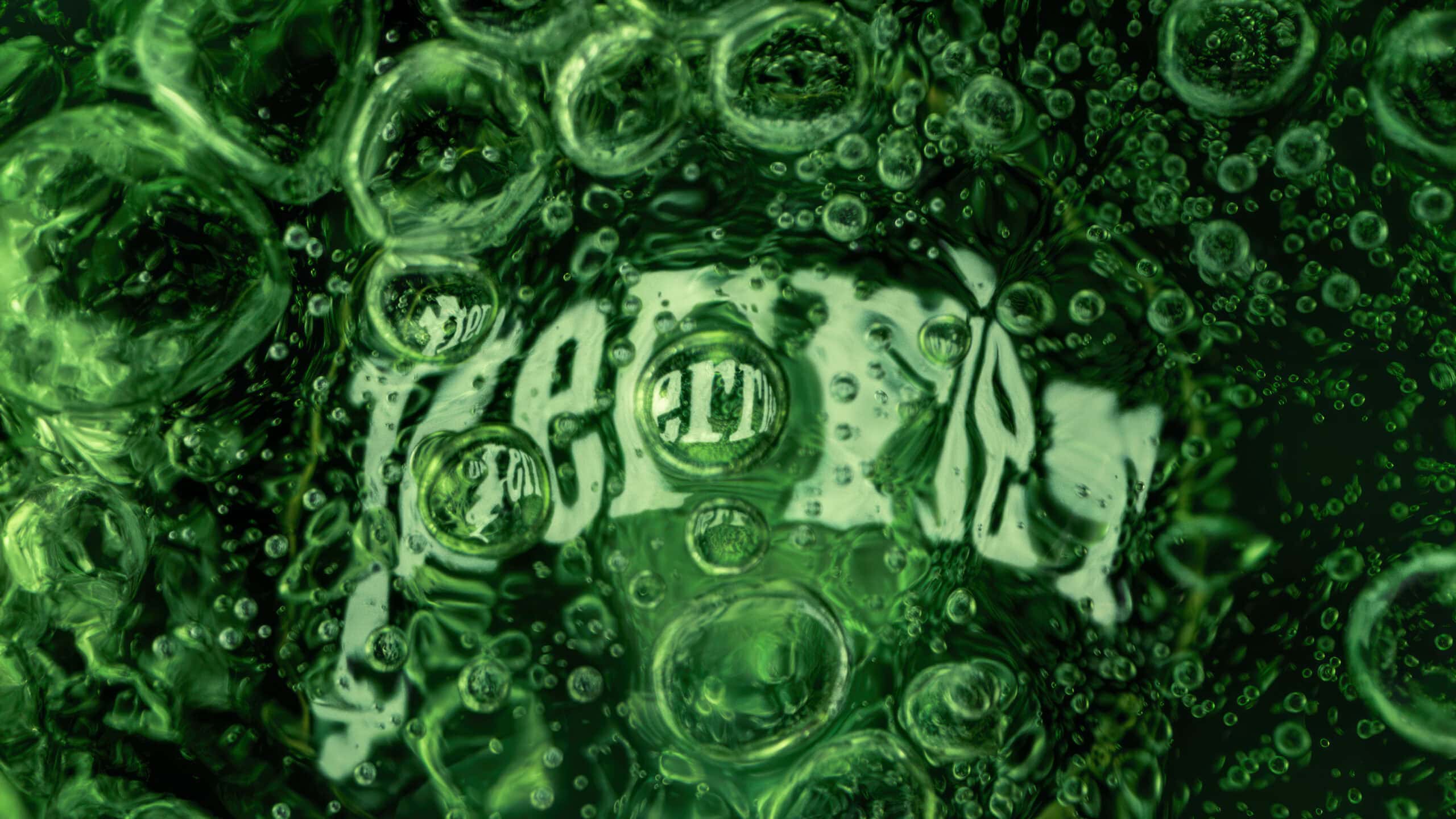 Macro image inside a green glass bottle of Perrier sparkling water. In the centre of the image you see the bottom of the glass with the word 'Perrier' written which has bubbles covering the logo.