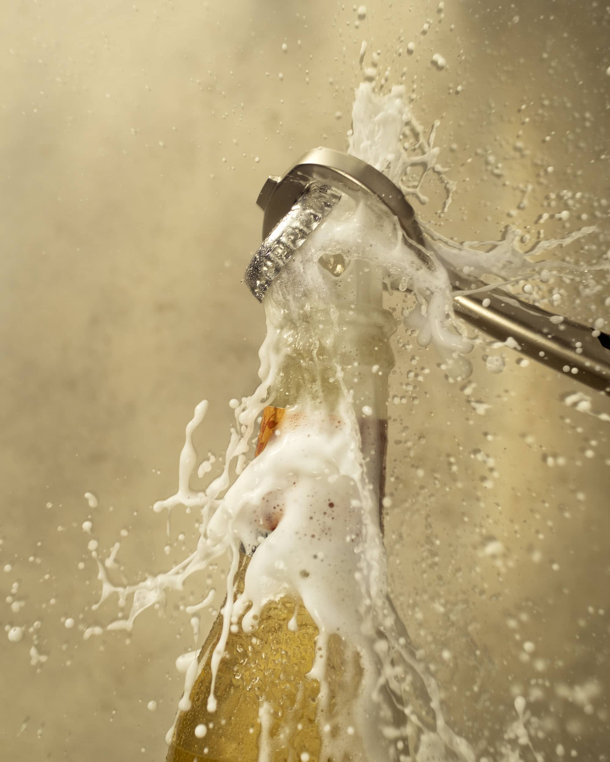 Close-up of a steel bottle opener as it takes the lid off a glass bottle of FeverTree ginger ale. The bottle cap is mid-air as fizzy liquid and foam bursts through the top of the bottle creating a big splash