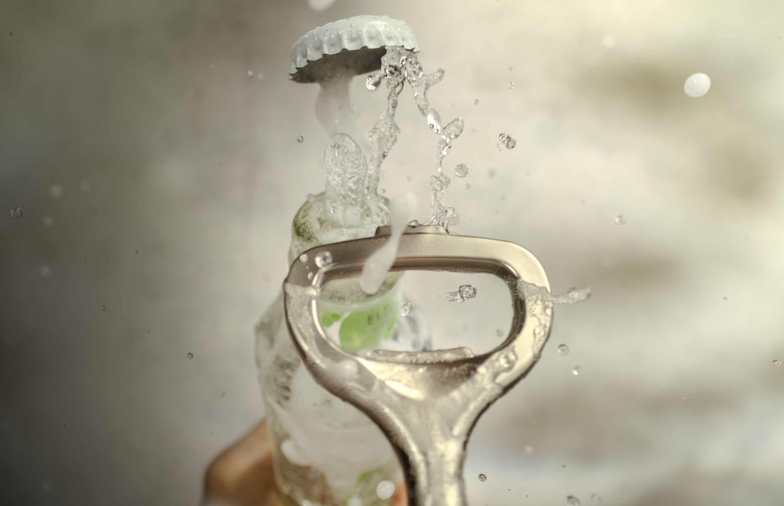 Close-up of a steel bottle opener as it takes the lid off a glass bottle of tonic water. Fizzy liquid and foam bursts through the top of the bottle creating a big splash