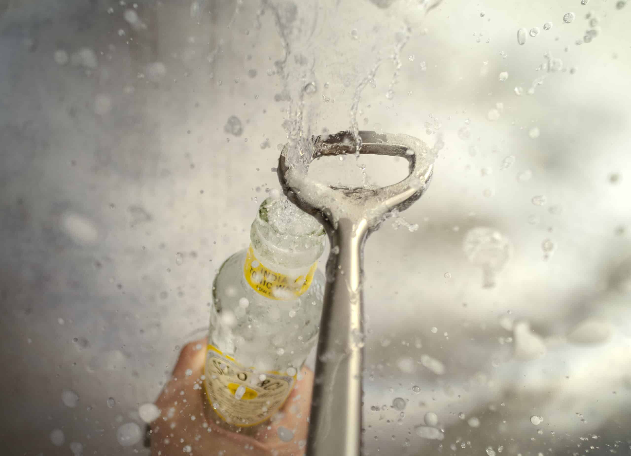 Close-up of a steel bottle opener as it takes the lid off a glass bottle of tonic water. Fizzy liquid and foam bursts through the top of the bottle creating a big splash