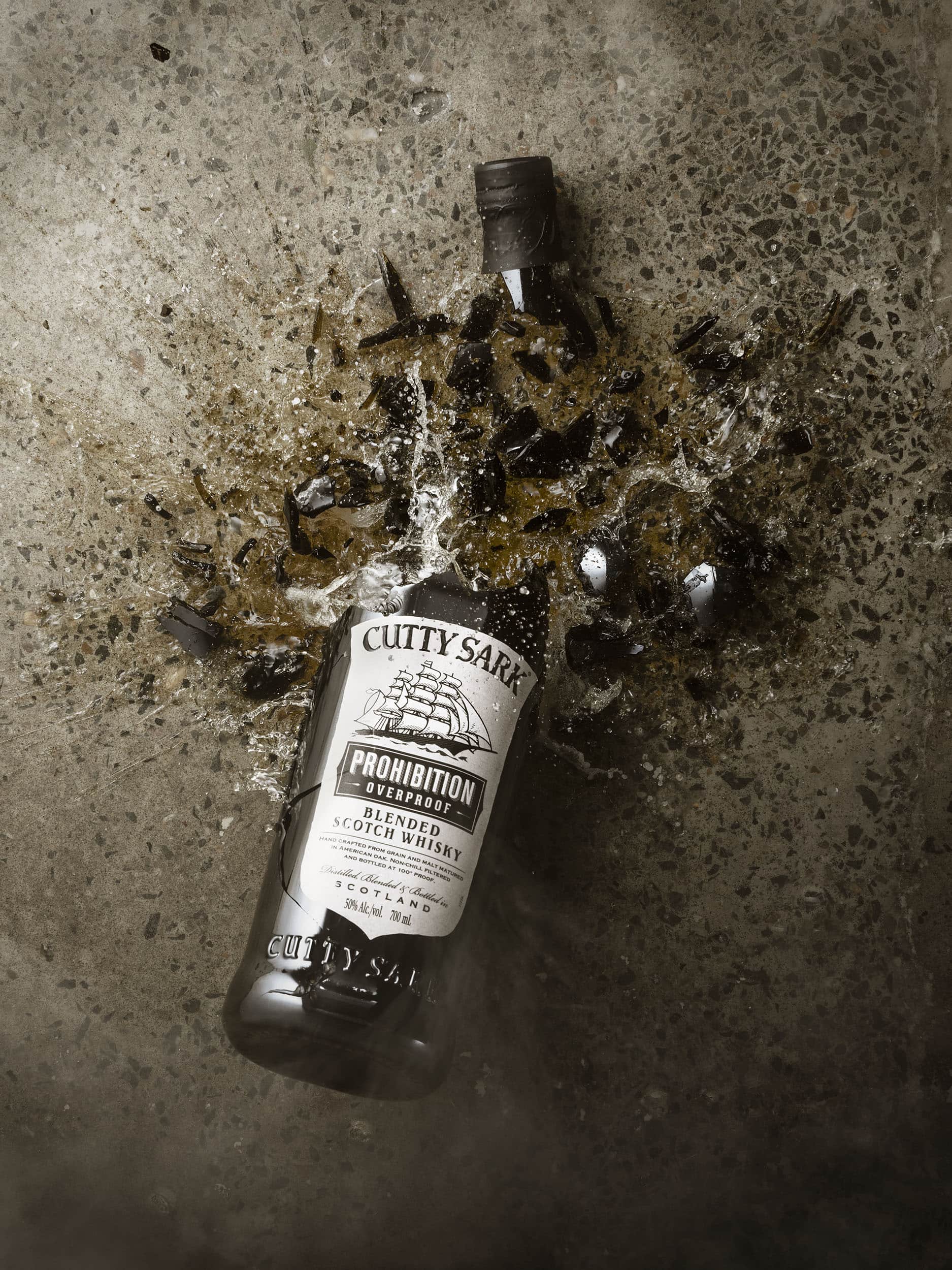 Action shot of a bottle of Cutty Sark Prohibition scotch whisky in mid-break as it smashes on a concreate floor. The bottom half of the bottle remains intact. The top of the bottle has smashed as you see orange liquid spilling our on to the floor.