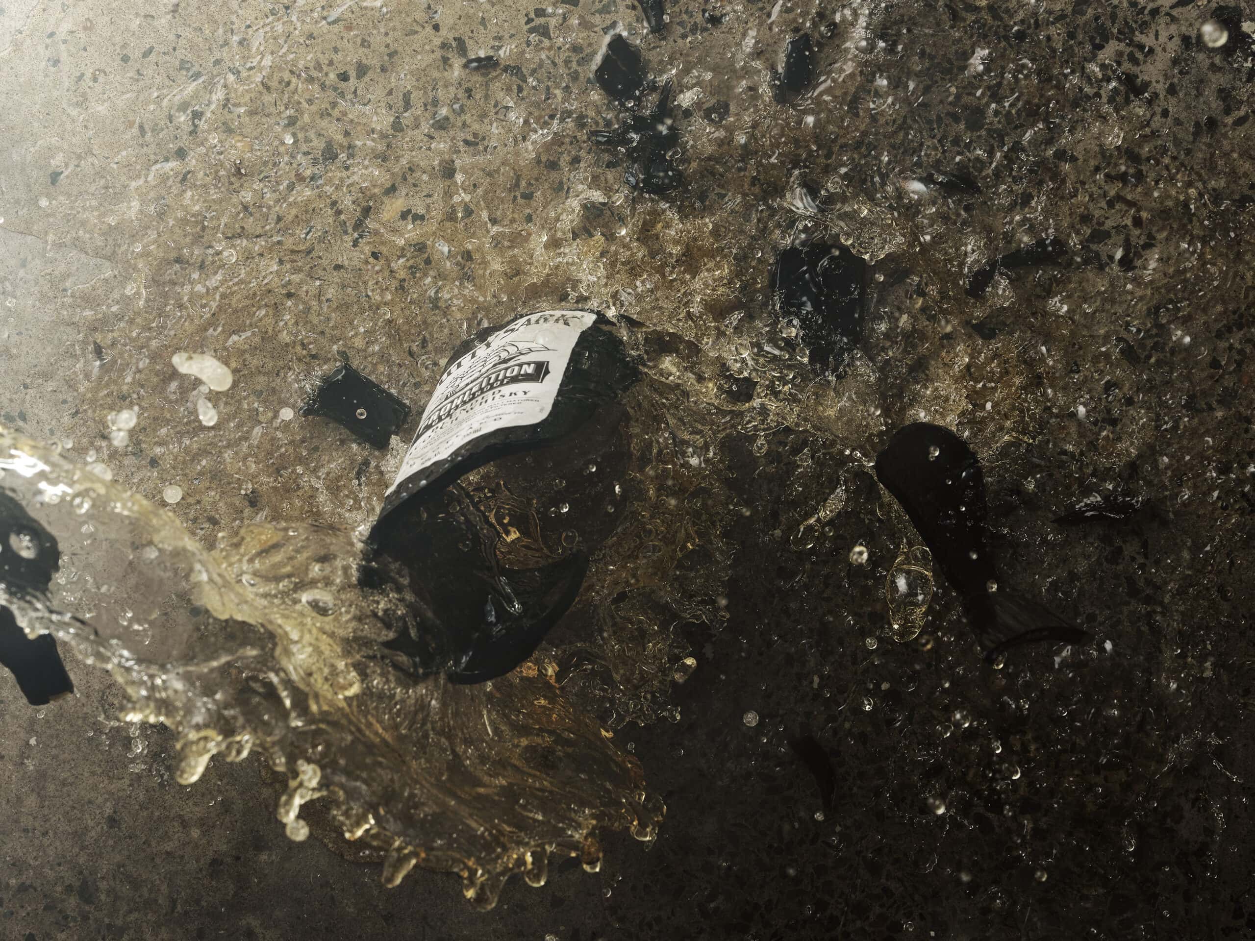 Action shot of a bottle of Cutty Sark Prohibition scotch whisky in mid-break as it smashes on a concreate floor. The bottom half of the bottle remains intact. The top of the bottle has smashed as you see orange liquid spilling our on to the floor.