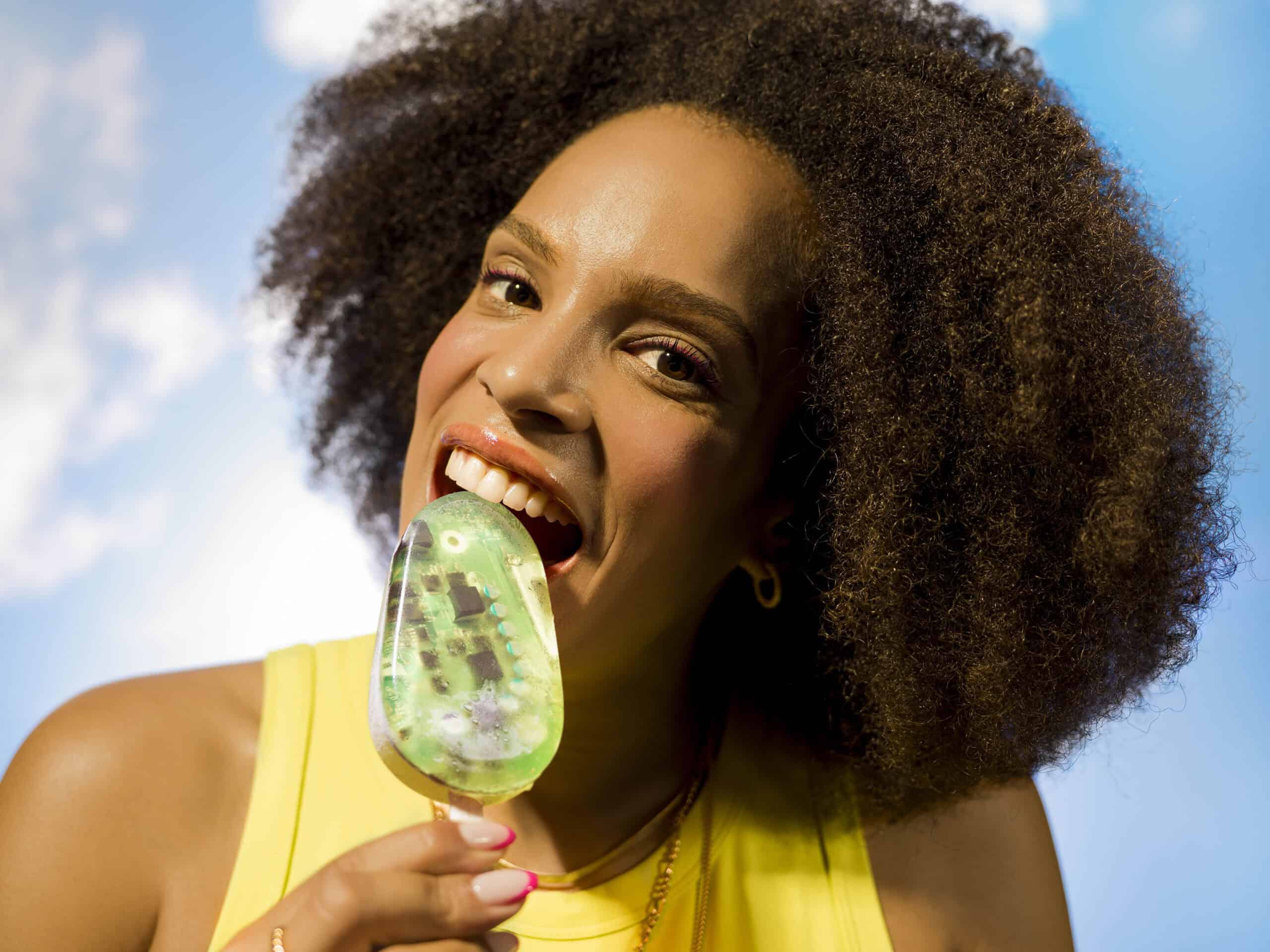 Black girl with afro hair about to bite into a green ice lolly that has tech chips placed inside. The background is a sunny blue sky