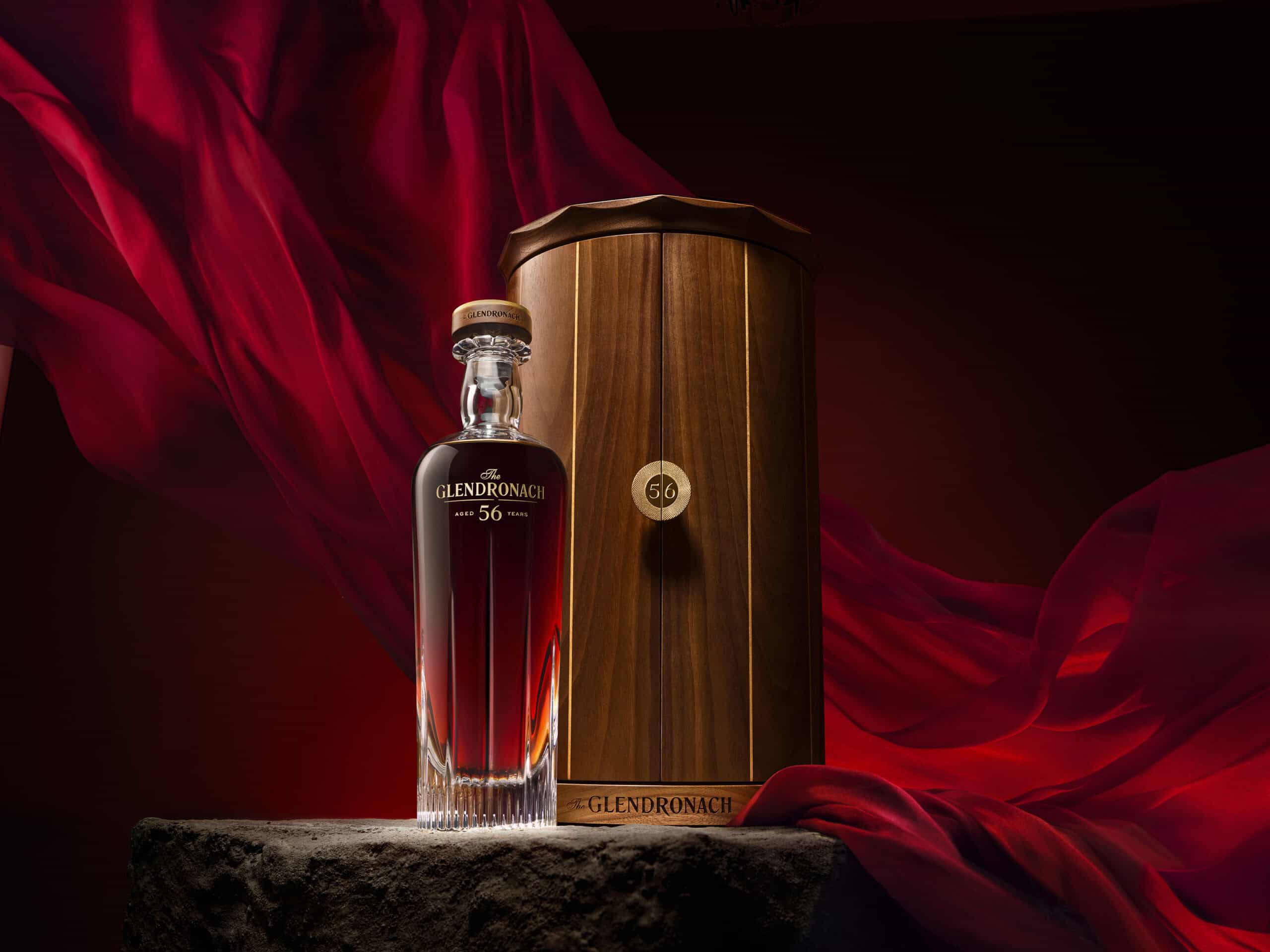 Whisky bottle sitting on a stone plinth with a Glendronach 56YO oak box next to it. The camera angel is low to make the bottle look heroic. There is a red drape in the background blowing in the wind.